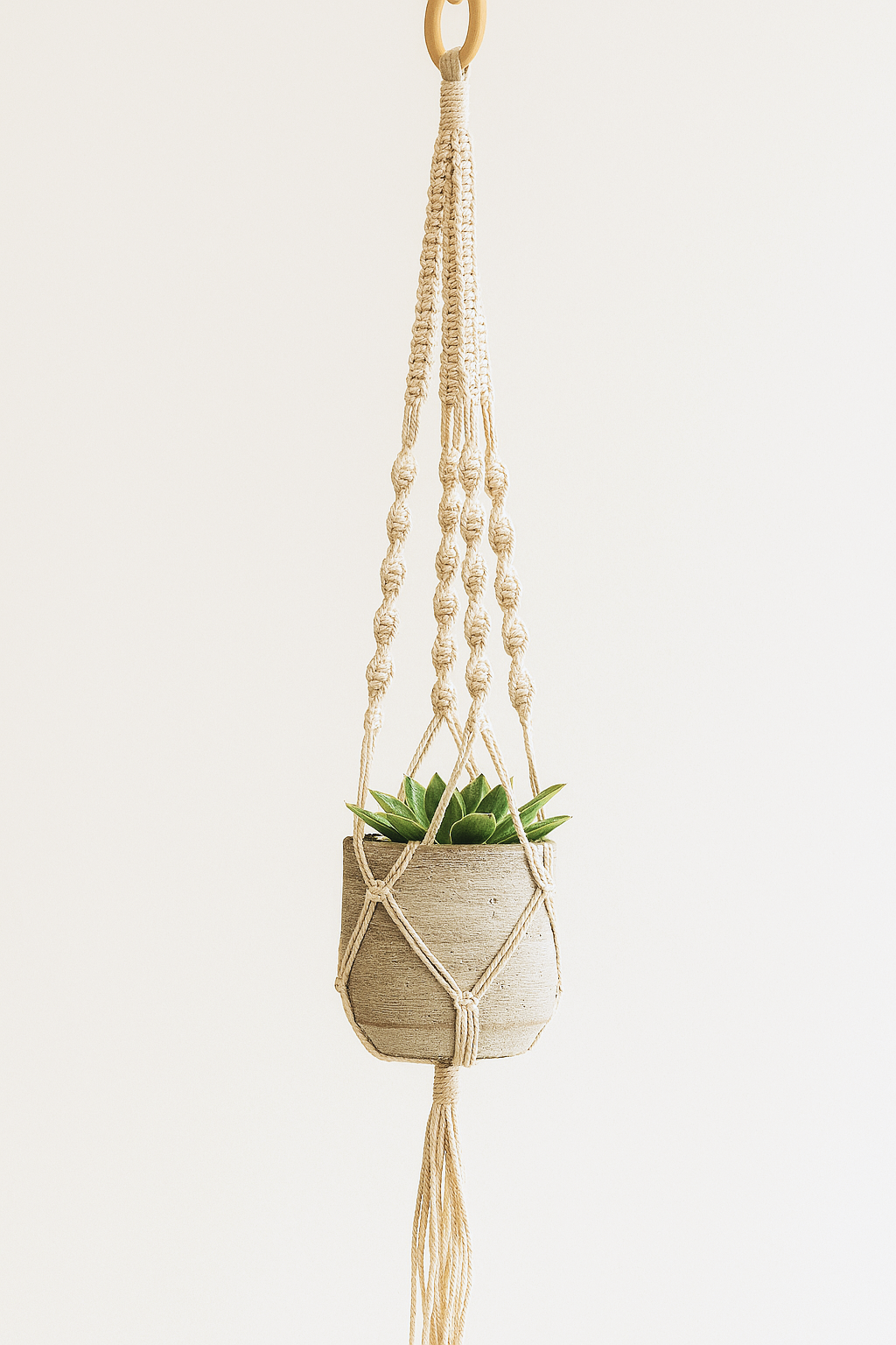 A handcrafted macramé plant hanger made from off-white cotton cord, featuring a small grey pot with a succulent plant. Styled against a warm cream background in a minimalist boho aesthetic.
