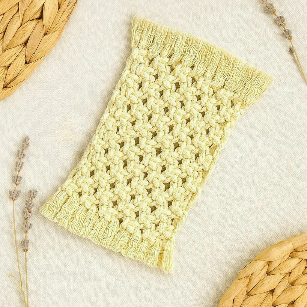 Flat lay of a handmade off white macramé mug rug by Samra Knots, styled with dried lavender sprigs and natural woven placemats on a neutral beige fabric background.