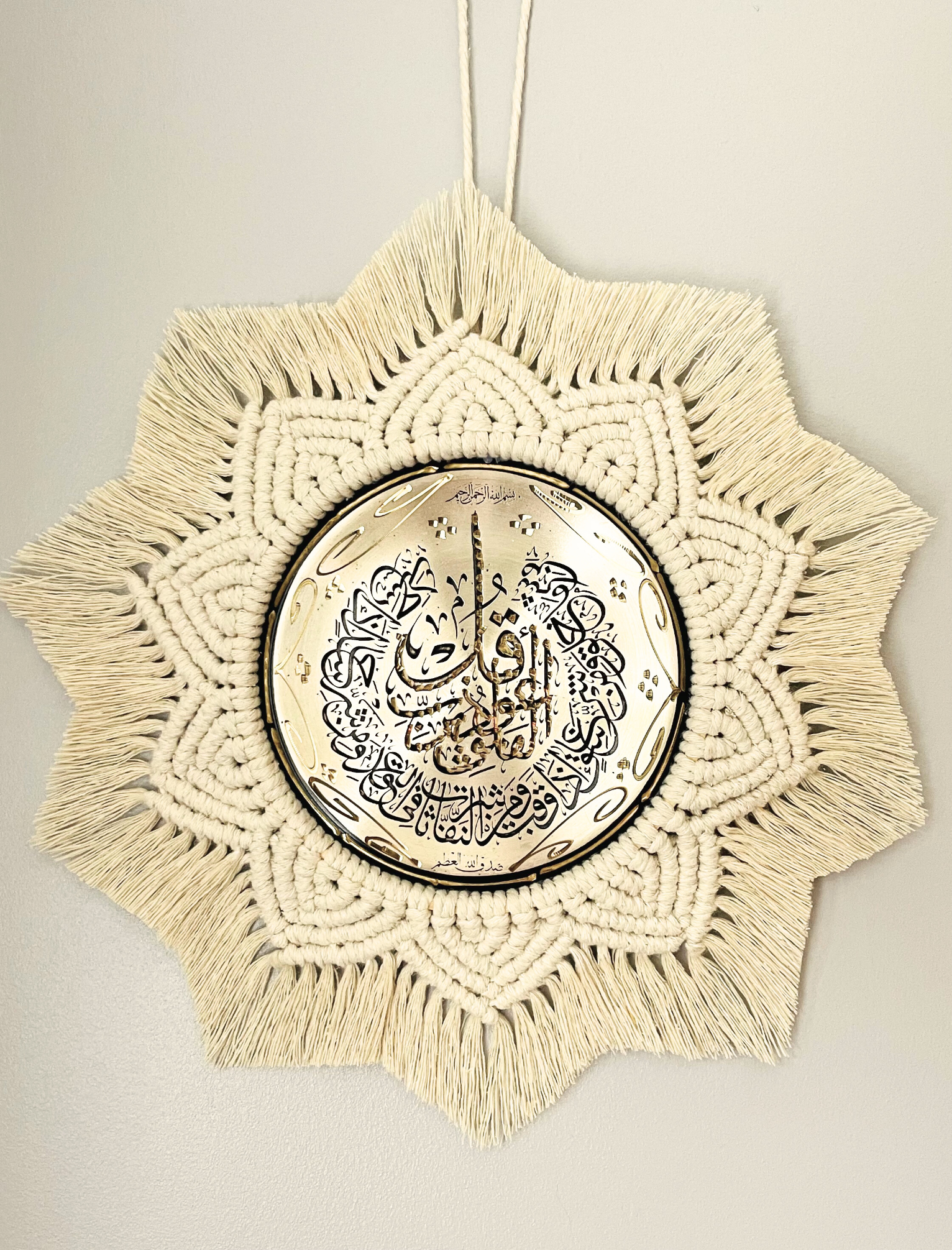 Handmade Islamic wall hanging featuring a gold metal plate with Arabic calligraphy, surrounded by intricate off-white macramé in a starburst pattern