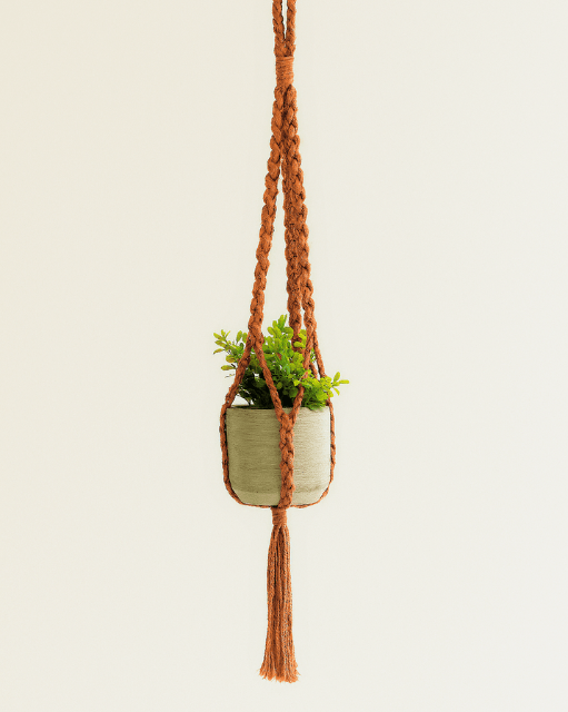 Handmade macramé plant hanger in terracotta, hanging on a light background with a concrete pot and green plant – boho home décor style
