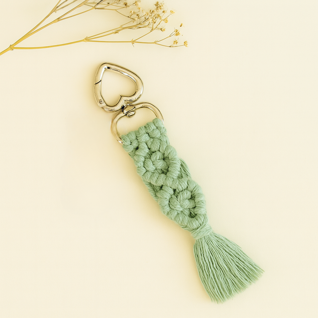 Soft sage green macramé keychain with silver heart clasp, displayed on a neutral background with a sprig of dried flowers