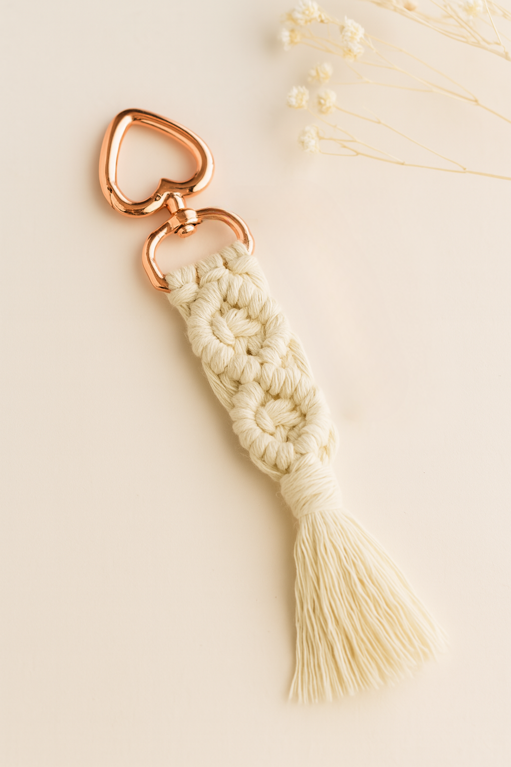Off-white handmade macramé keychain with rose gold heart clasp, laid on a soft neutral background with dried baby's breath