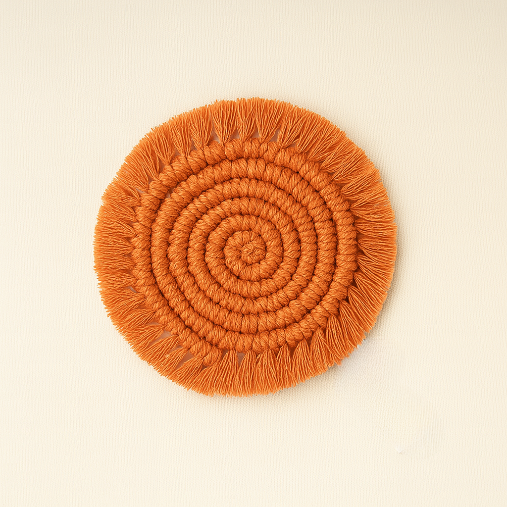 A handcrafted round macramé coaster in a warm terracotta color with a spiral design and fringe edges, lying flat on a beige textile background with a "Samra Knots" label.