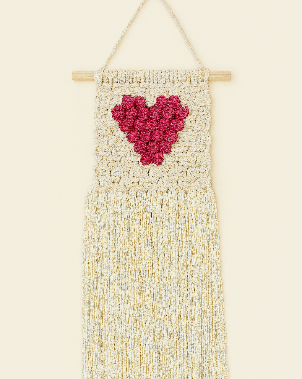 A handmade macramé wall hanging with a raised pink heart on an off-white cotton background and fringe, hanging from a wooden dowel.