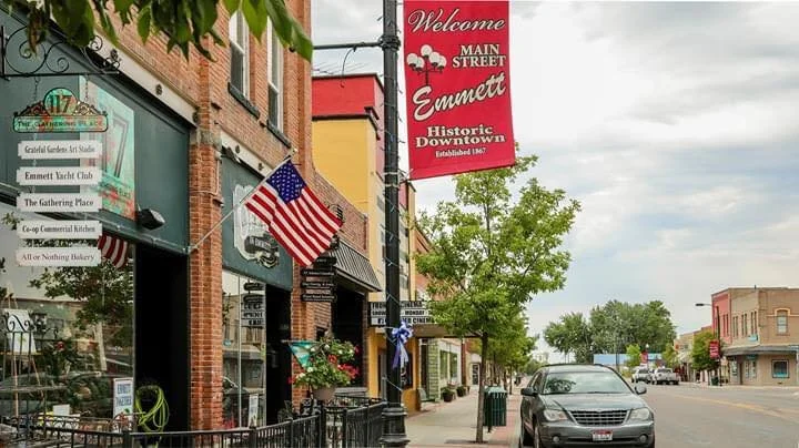 Main Street in Emmett, Idaho, with businesses and storefronts, American flags, a red Welcome to Emmett sign, and a gray car parked along the sidewalk under an overcast sky.