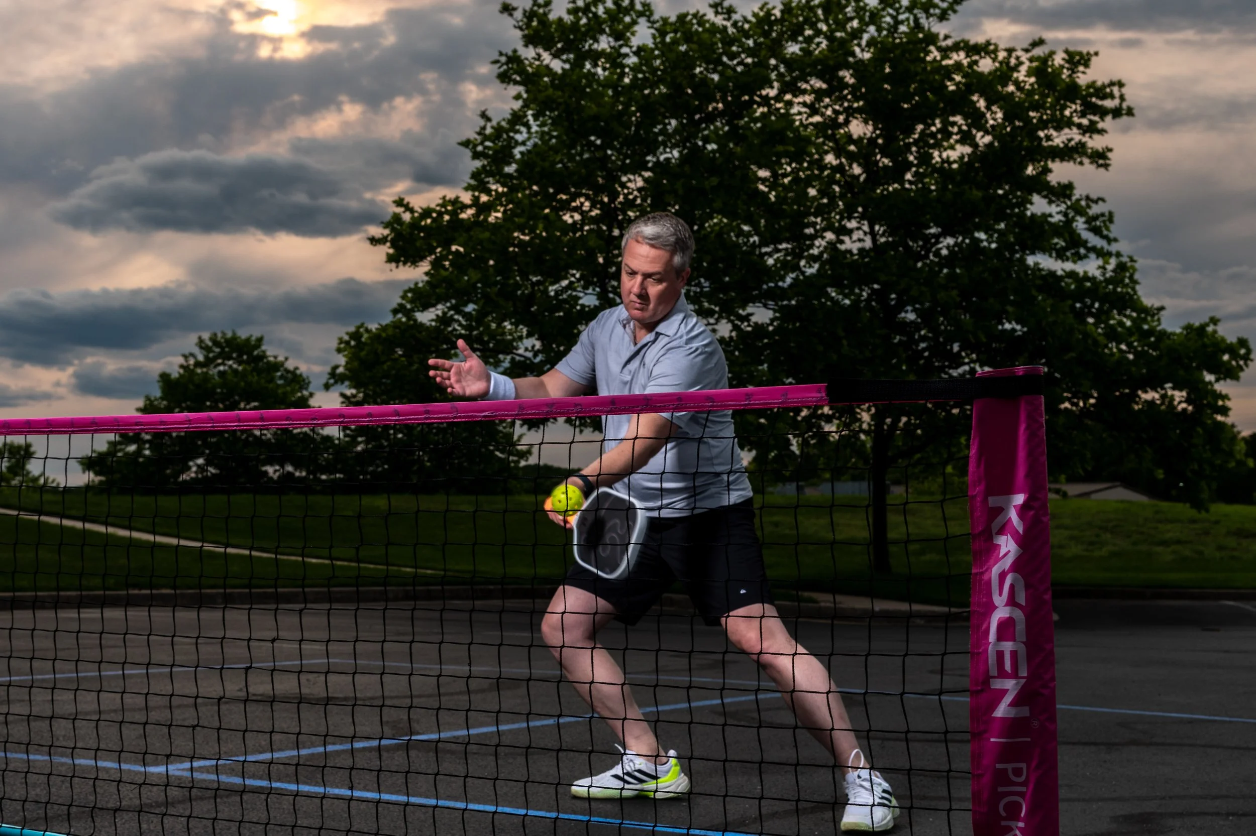 A man playing pickleball on an outdoor court at dusk, holding a yellow pickleball and preparing to hit it with a paddle, with trees and a cloudy sky in the background.