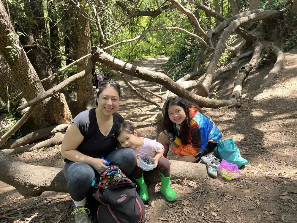 A parent and two children resting on a tree root on a shaded trail in the East Bay.