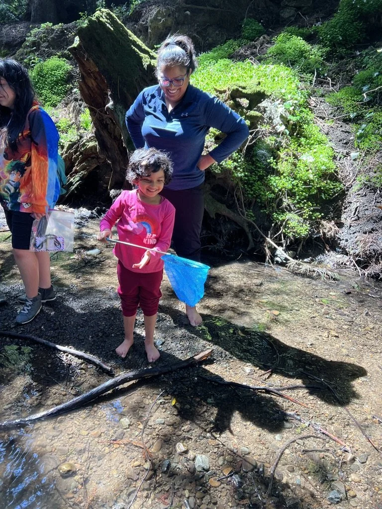 A child holding a butterfly net in a shallow creek beside a parent in the East Bay.