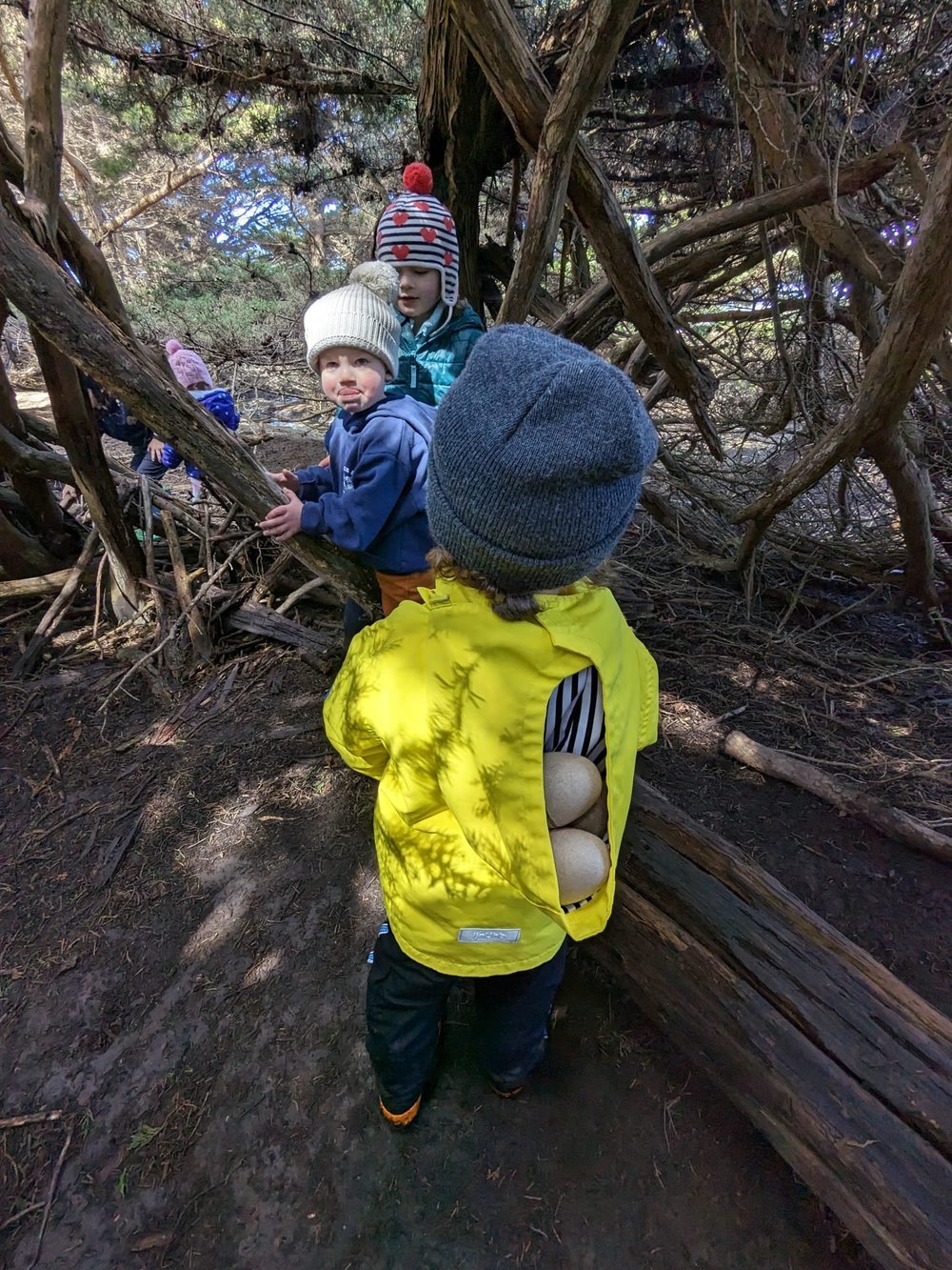 Young children in hats exploring among twisted cypress branches, one with eggs in a jacket pocket.