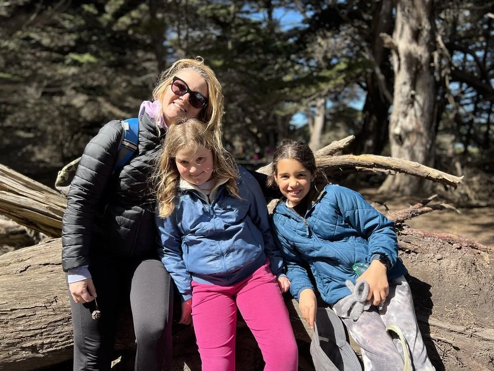 A parent and two children smiling on a fallen log among cypress trees.