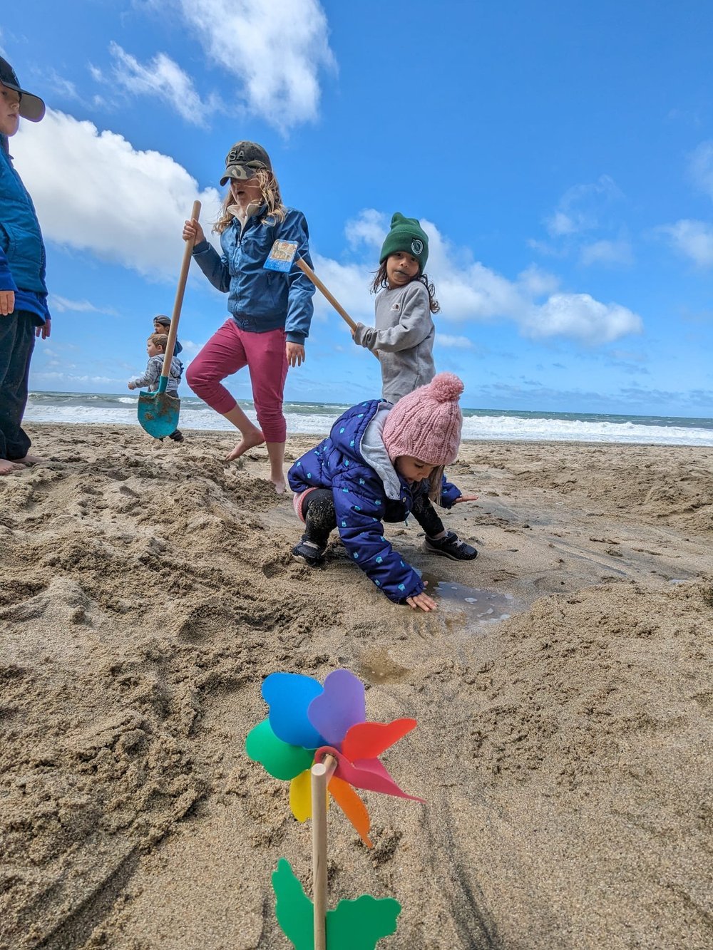 Children digging in the sand on a beach, with a colorful pinwheel in the foreground.