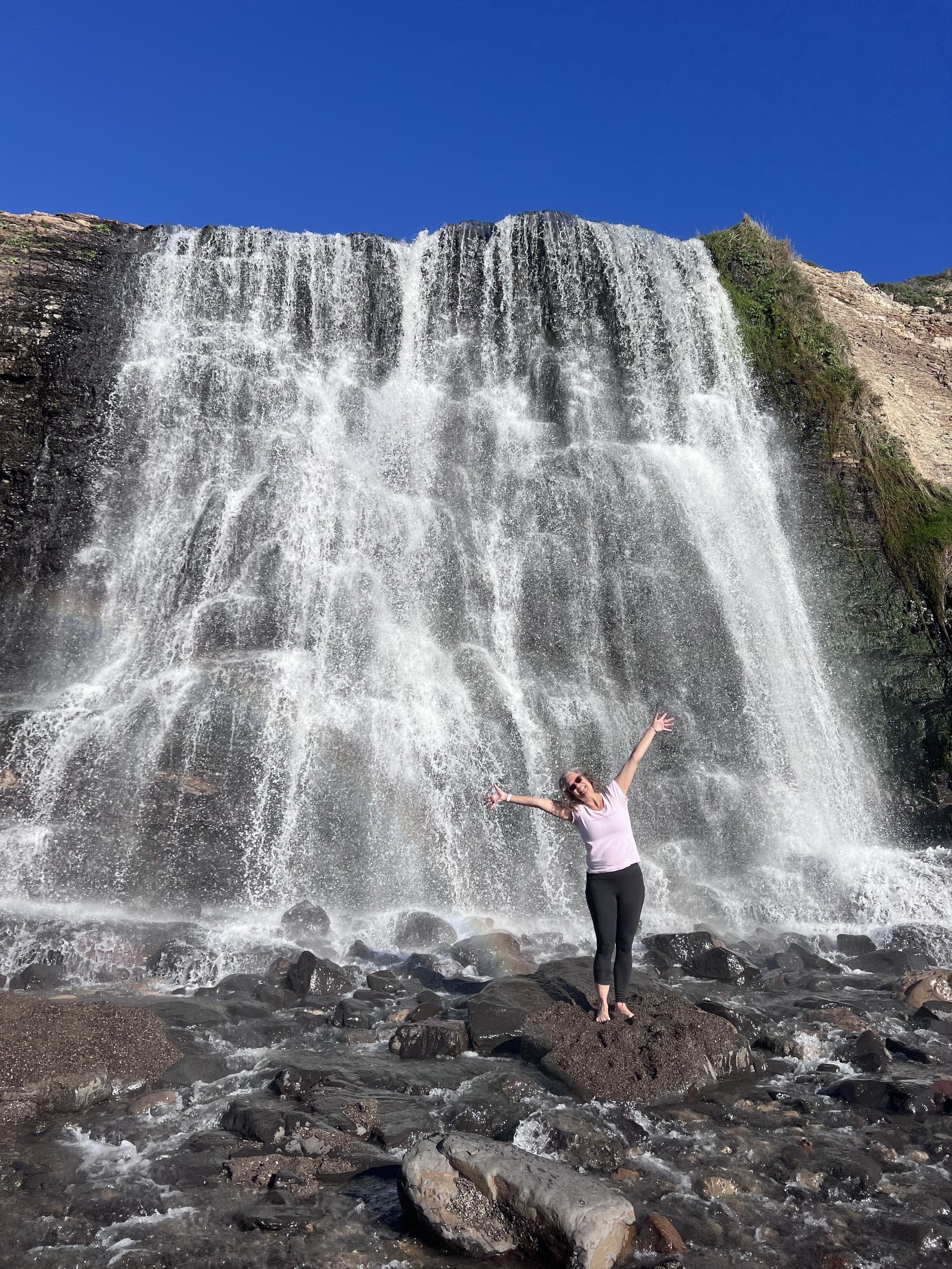 A woman with her arms raised at the foot of Alamere Falls, Point Reyes.