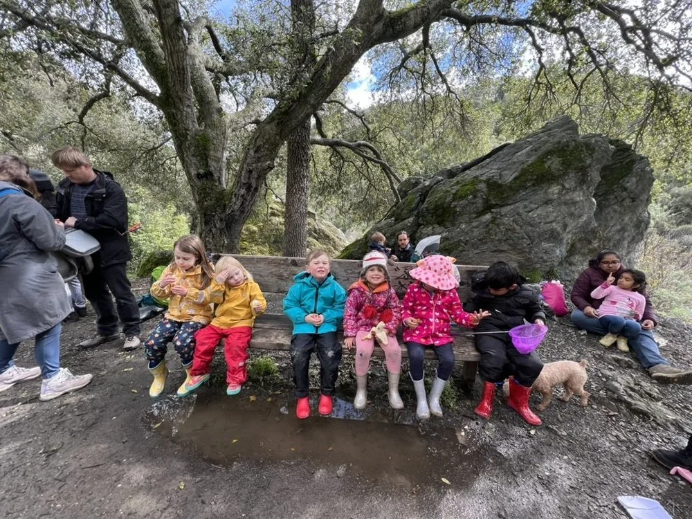 A row of children in rain boots sitting on a bench under an oak at Little Yosemite in the East Bay.