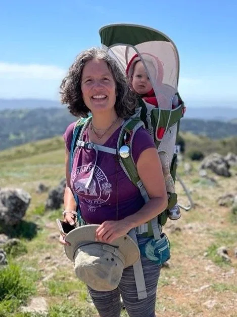 A smiling woman on a hilltop wearing an Osprey Poco backpack carrier with a baby under its sunshade.