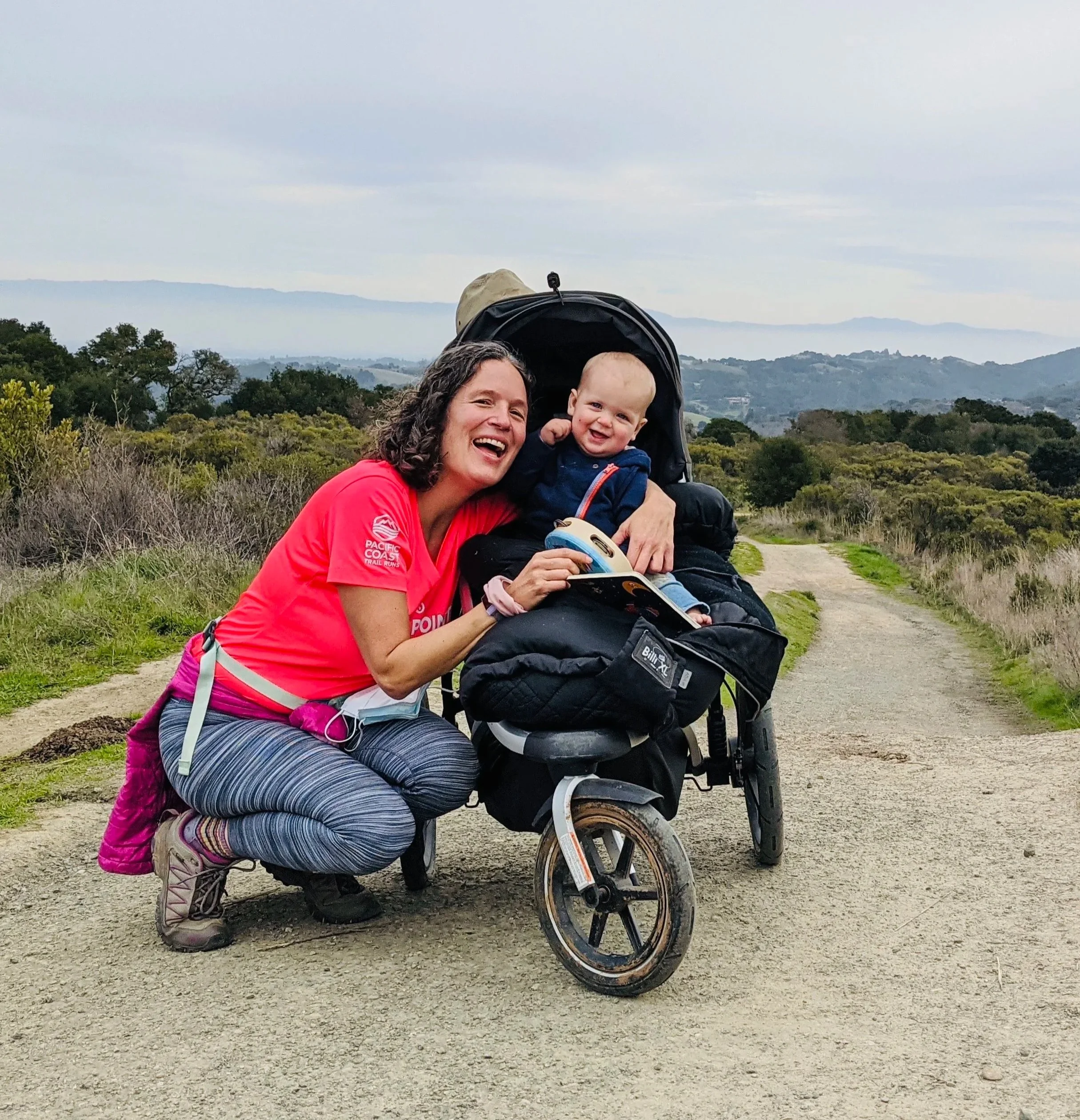 A smiling mother kneeling beside her laughing baby in a Thule jogging stroller on a hilltop dirt trail.