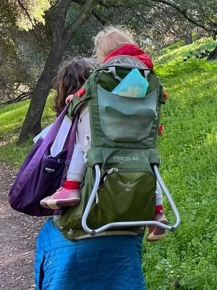 An adult walking a woodland trail with a child in a green Osprey Poco backpack carrier, seen from behind.