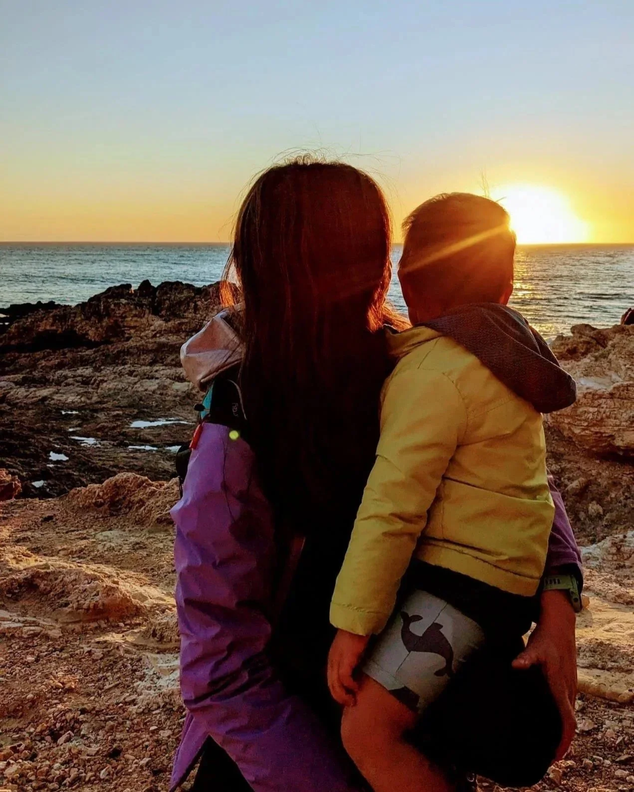 A woman and a young boy standing on rocky terrain by the ocean, watching a sunset.