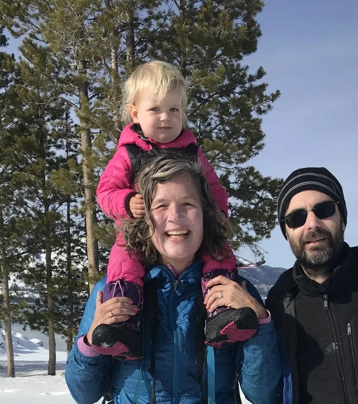 A toddler in a pink puffy suit riding on a smiling woman's shoulders, a man beside them and snowy pines behind.