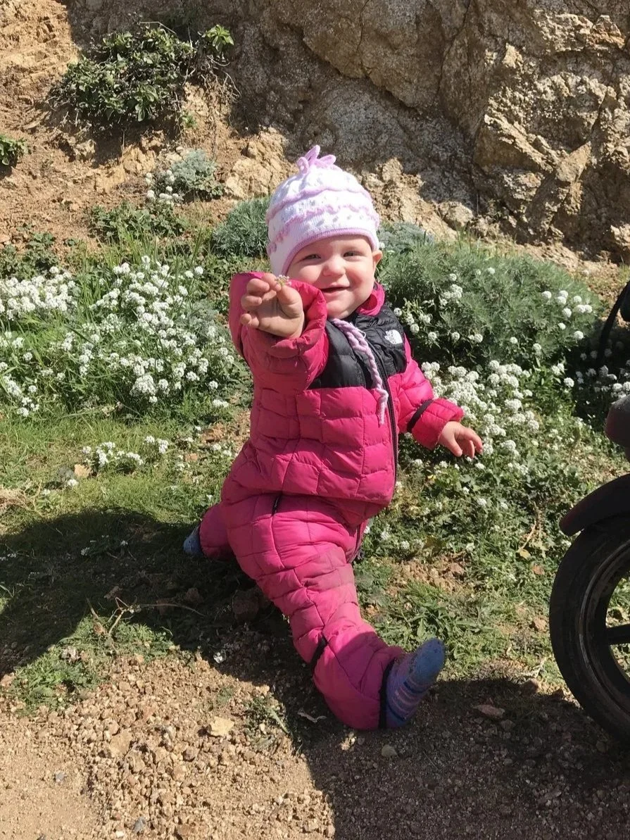 A toddler in a pink The North Face ThermoBall suit and knit hat sitting by white wildflowers.