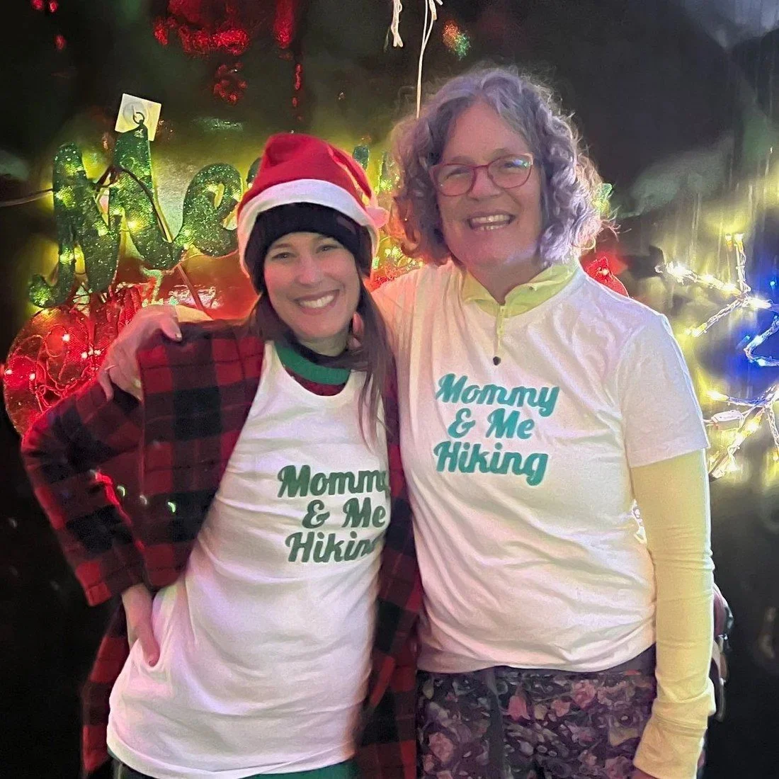 Dani and Miranda of Family Adventure Squad in matching Mommy & Me Hiking T-shirts, smiling in front of holiday lights