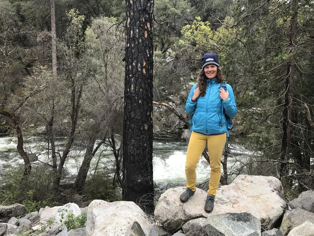 Mom in a blue down jacket and beanie standing on granite boulders beside a rushing river in a pine forest, dressed warm for winter camping