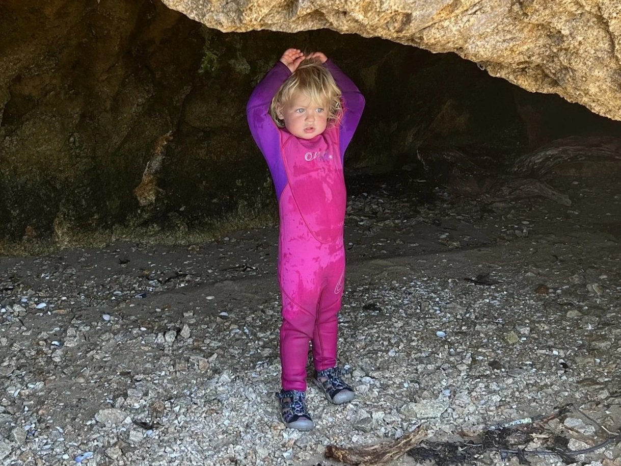 A toddler in a pink O'Neill wetsuit standing at the mouth of a rocky sea cave on a pebble beach.