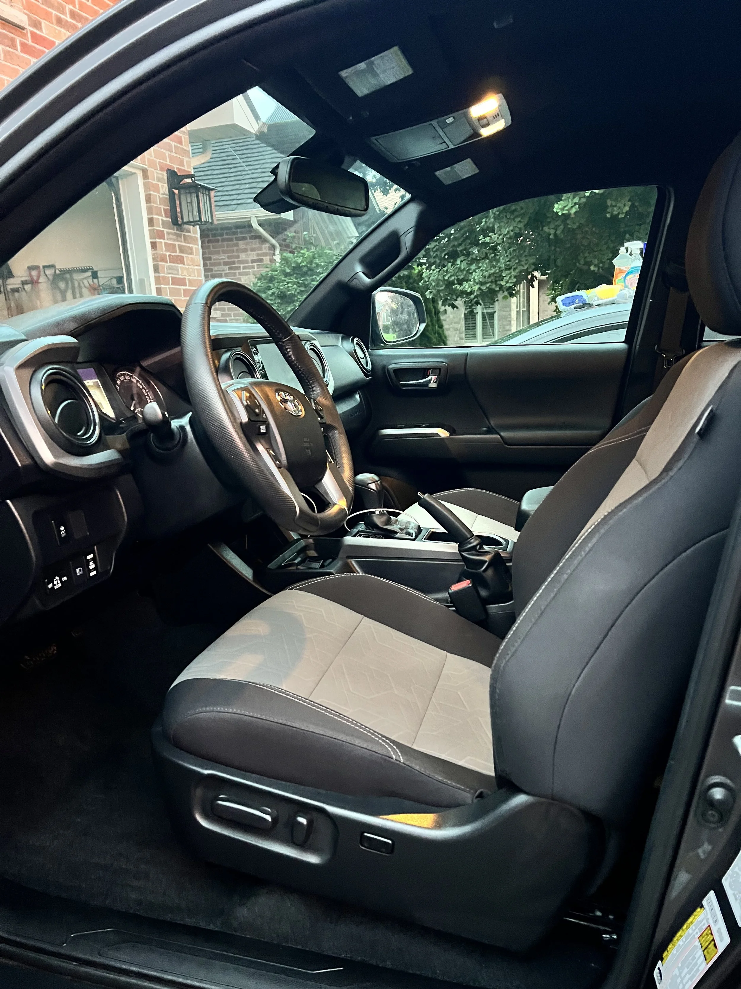 The image shows the interior of a vehicle, focusing on the front passenger seat and driver's area. The dashboard, steering wheel, and center console are visible, with a view of the outside through the window including a brick house and green foliage.