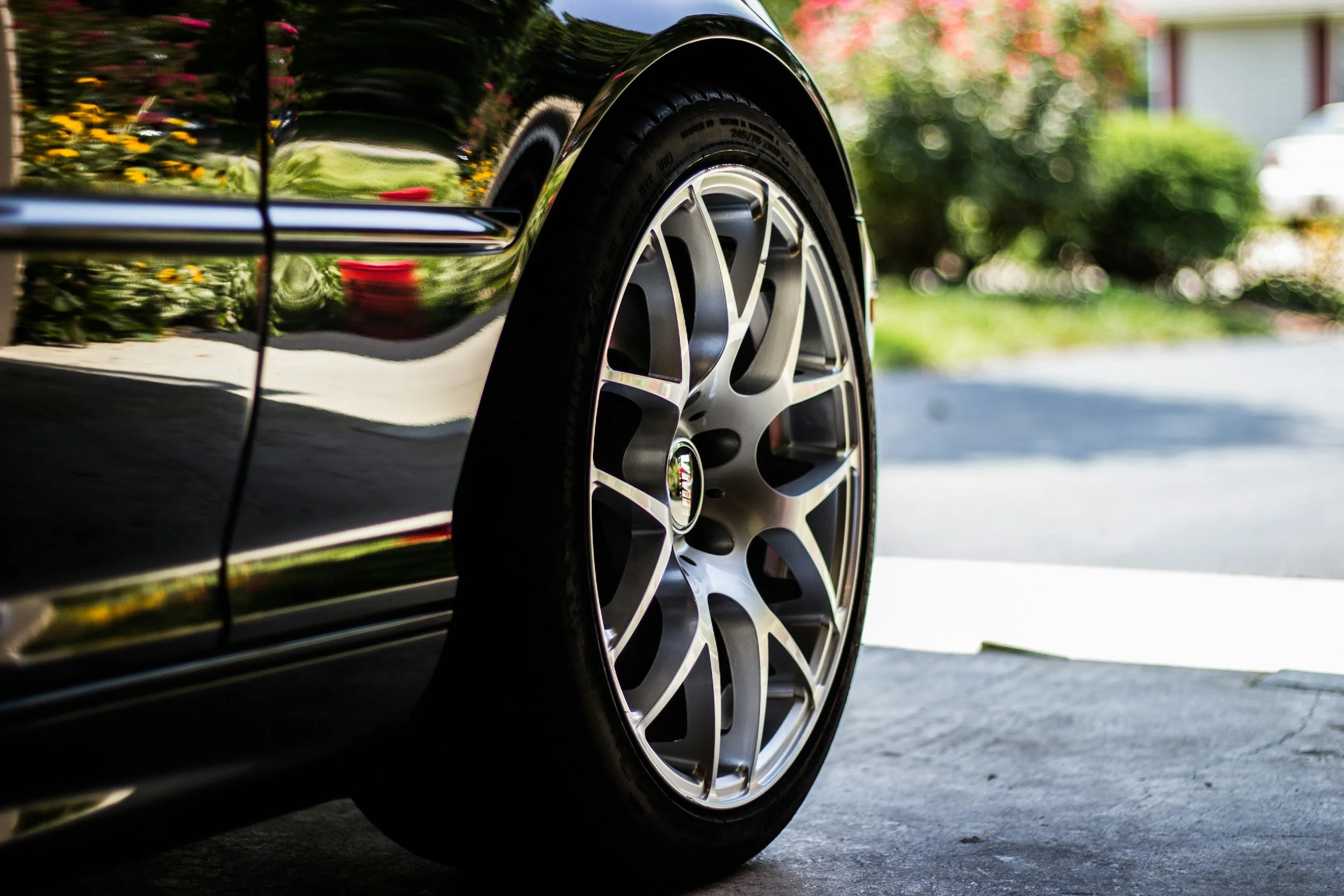 Close-up of a black sports car's front wheel with a chrome rim, parked outside on a driveway, with reflections of green bushes and a house on its surface.