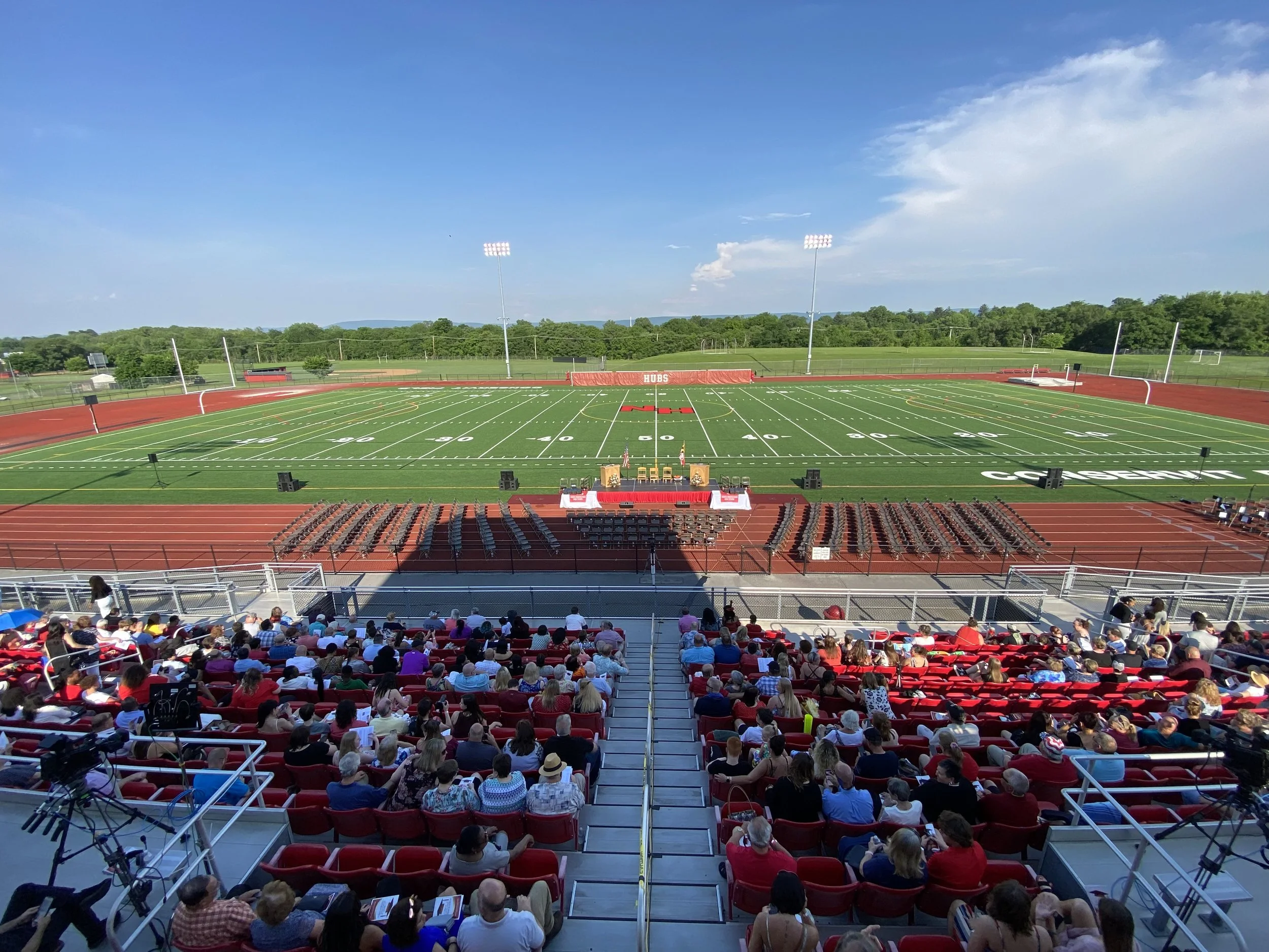 Photo of a crowded sports stadium with a football field, audience seated in red chairs, and a stage set up at the front with chairs and banners, under a clear blue sky.