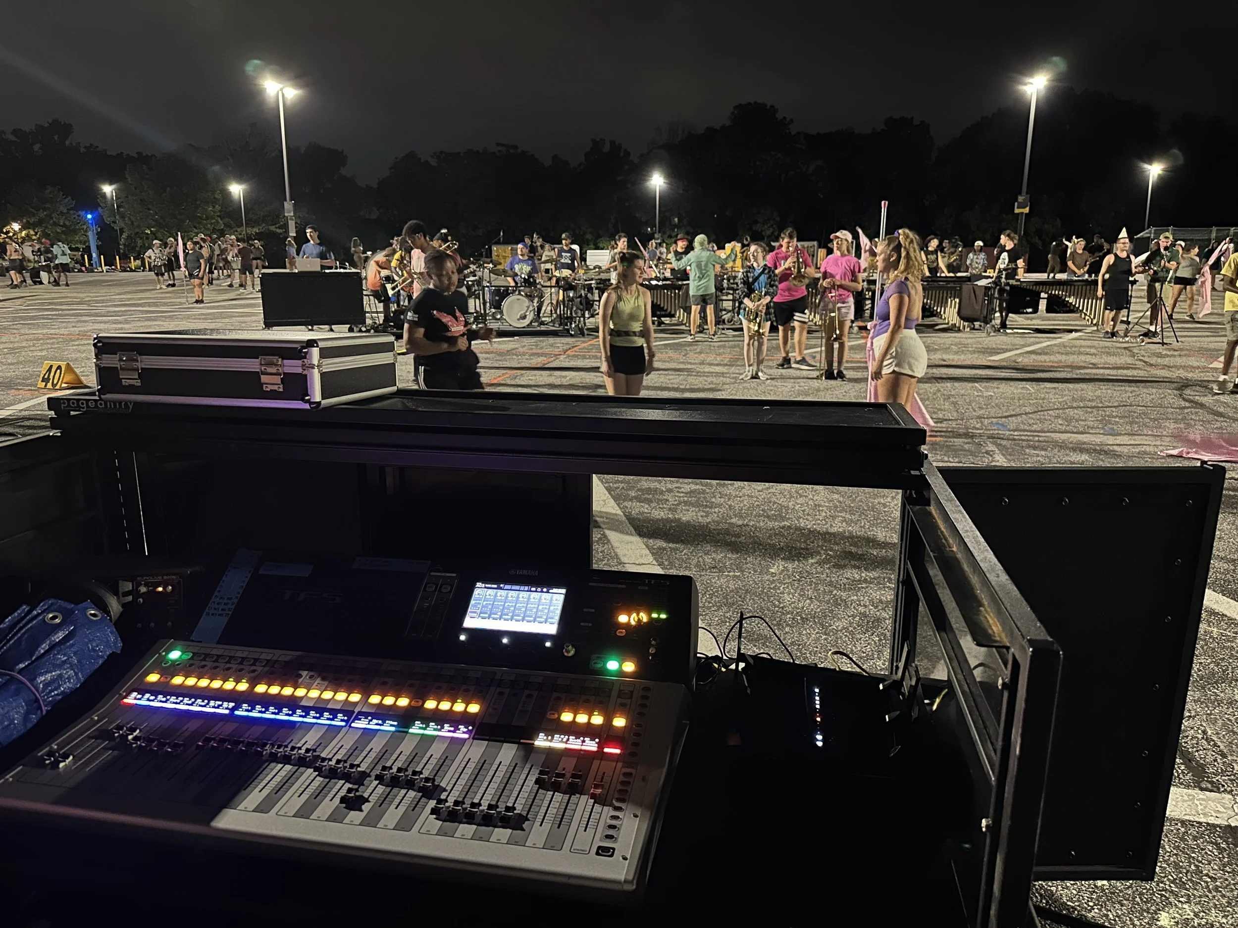 View of a parking lot at night during a live outdoor music event with musical instruments, people dancing, and a sound mixing console in the foreground.