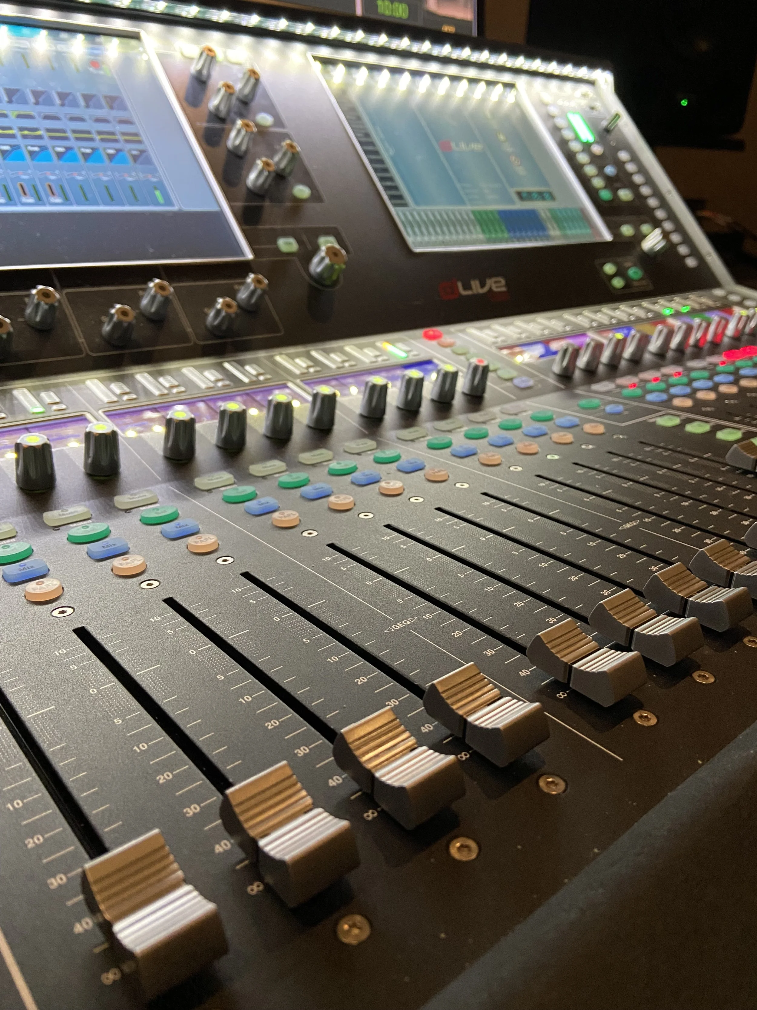 Close-up of a professional audio mixing console with multiple faders, knobs, and touchscreen displays.