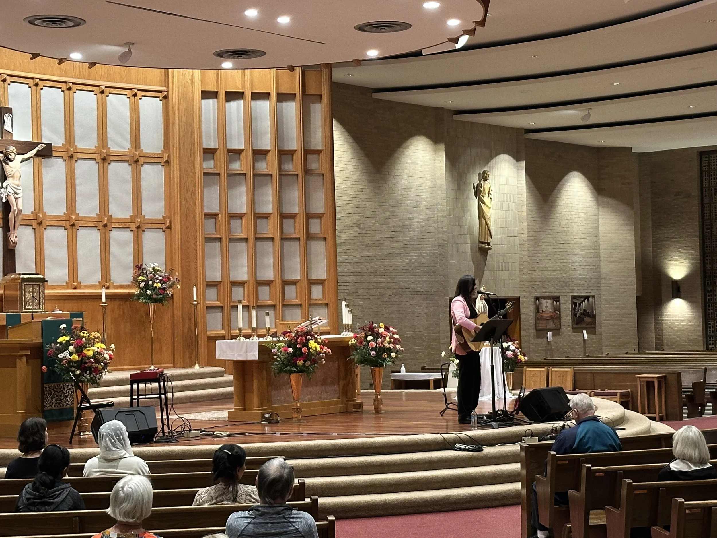 A woman playing guitar and singing at the front of a church with audience members seated in pews.
