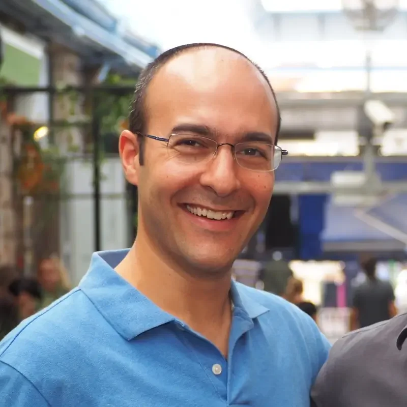A smiling man, Yoni Sherizen, CEO of Gabriel Protects, with short dark hair, wearing glasses and a blue polo shirt, standing indoors with a blurred background of people and structures.