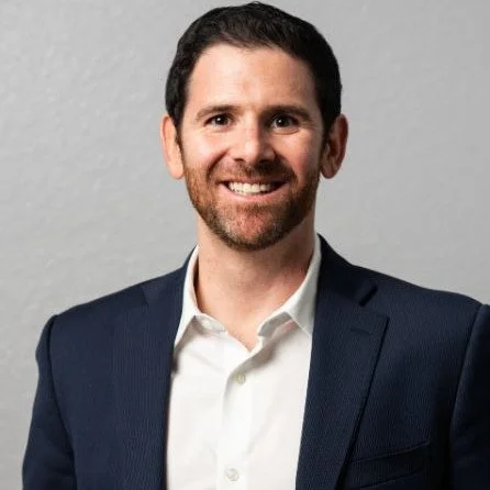 Smiling man in a navy suit and white shirt on gray background.