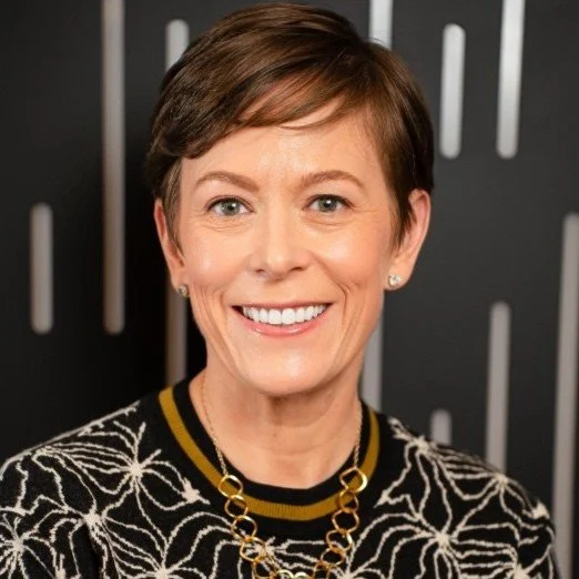 Portrait of a smiling woman with short brown hair, wearing a patterned black and white top and a gold necklace, in front of a dark background with vertical white lines.