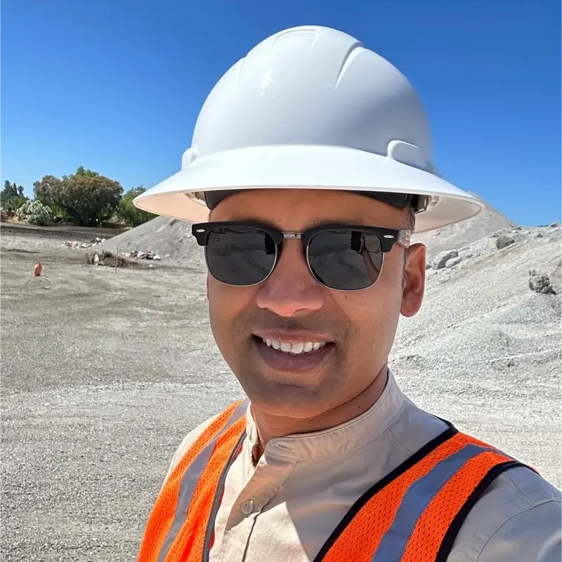 A man, Amar Amte (Founder and CEO at pegbo), at a construction site wearing a white hard hat, black sunglasses, and an orange safety vest. The background shows a gravelly area under a clear blue sky.