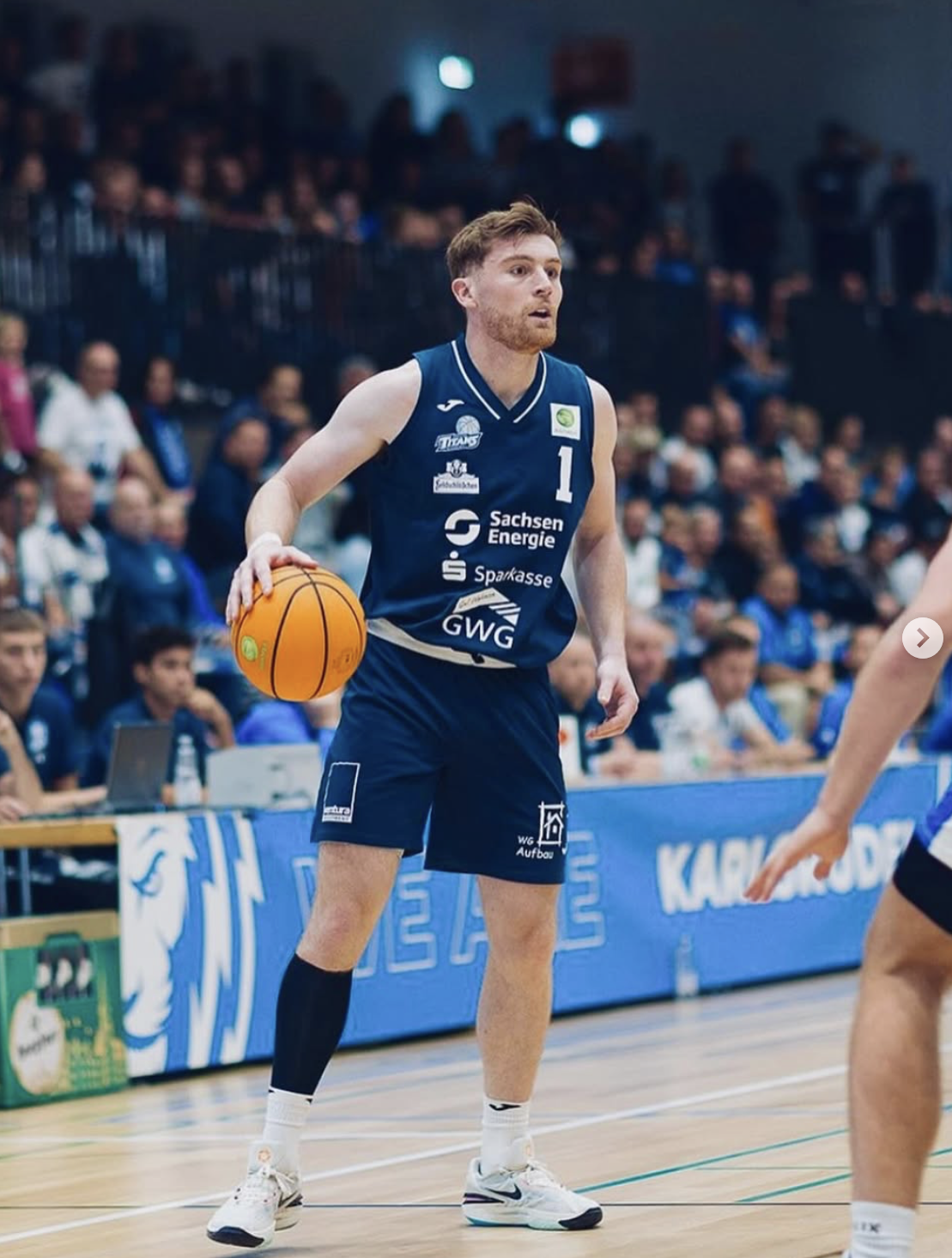 A male basketball player in a blue jersey holding an orange basketball on a court during a game, with a crowd in the background.