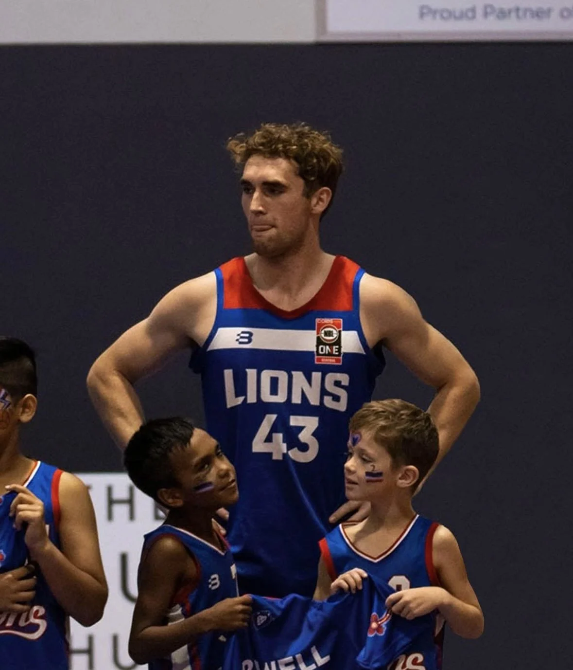 A male basketball player wearing a blue jersey with 'LIONS 43' standing with two young boys, one black and one white, wearing matching team jerseys and face paint, on a basketball court.