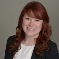 Smiling person with long red hair wearing a black blazer and white shirt against a neutral background.