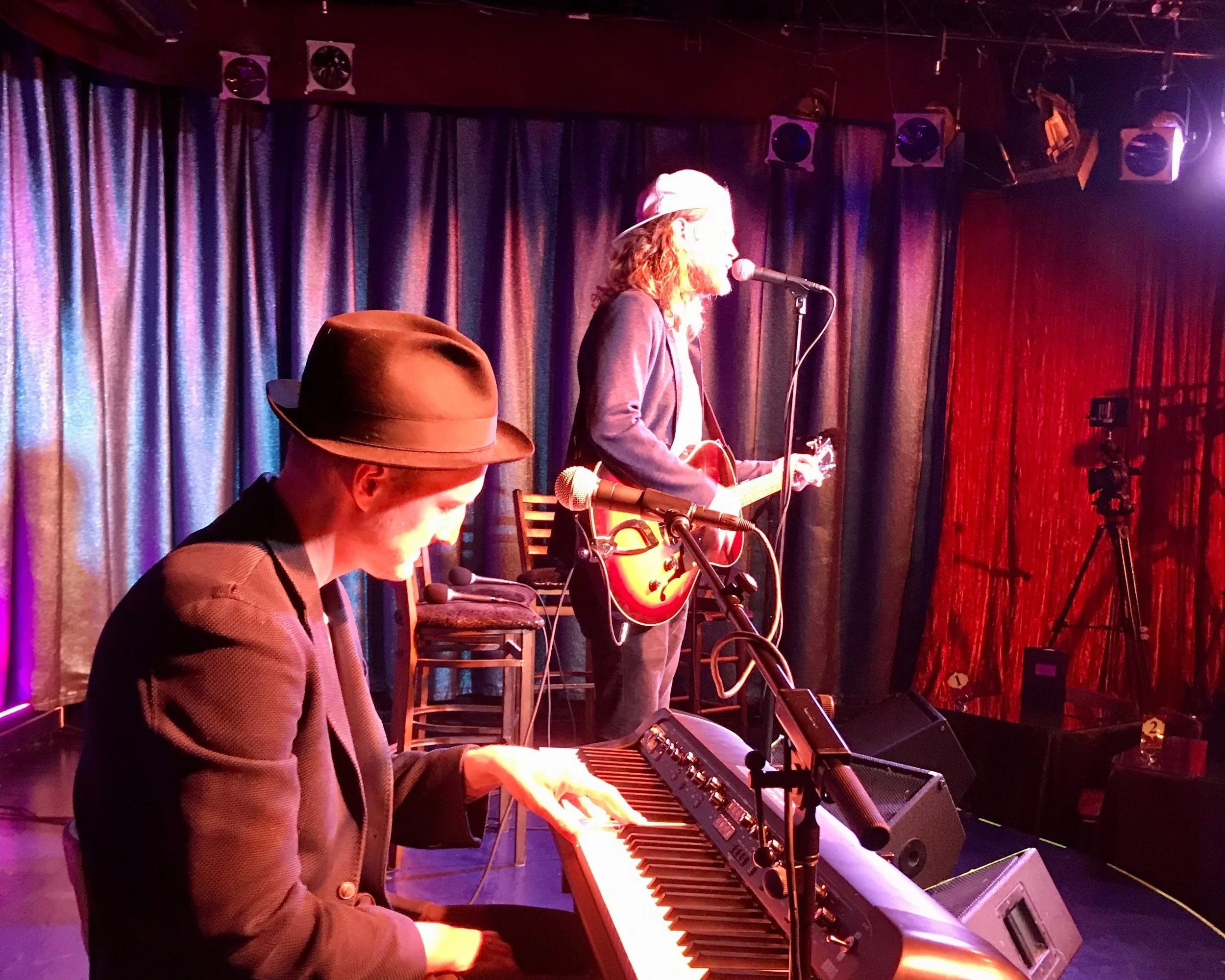 Two musicians performing on stage, one playing a keyboard and wearing a hat, the other singing and playing an electric guitar, in a dimly lit venue with curtains in the background.