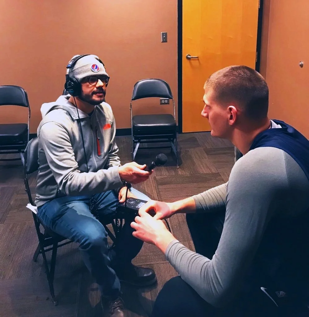 A man wearing a beanie and headphones is conducting an interview, holding a microphone towards another man who is seated across from him. The setting appears to be a small room with folding chairs and a closed door. Both men are casually dressed.