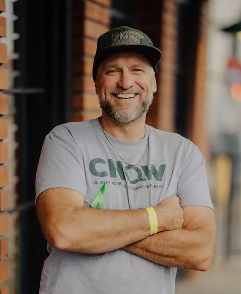 Man in gray T-shirt and cap smiling with arms crossed, standing near a brick wall.