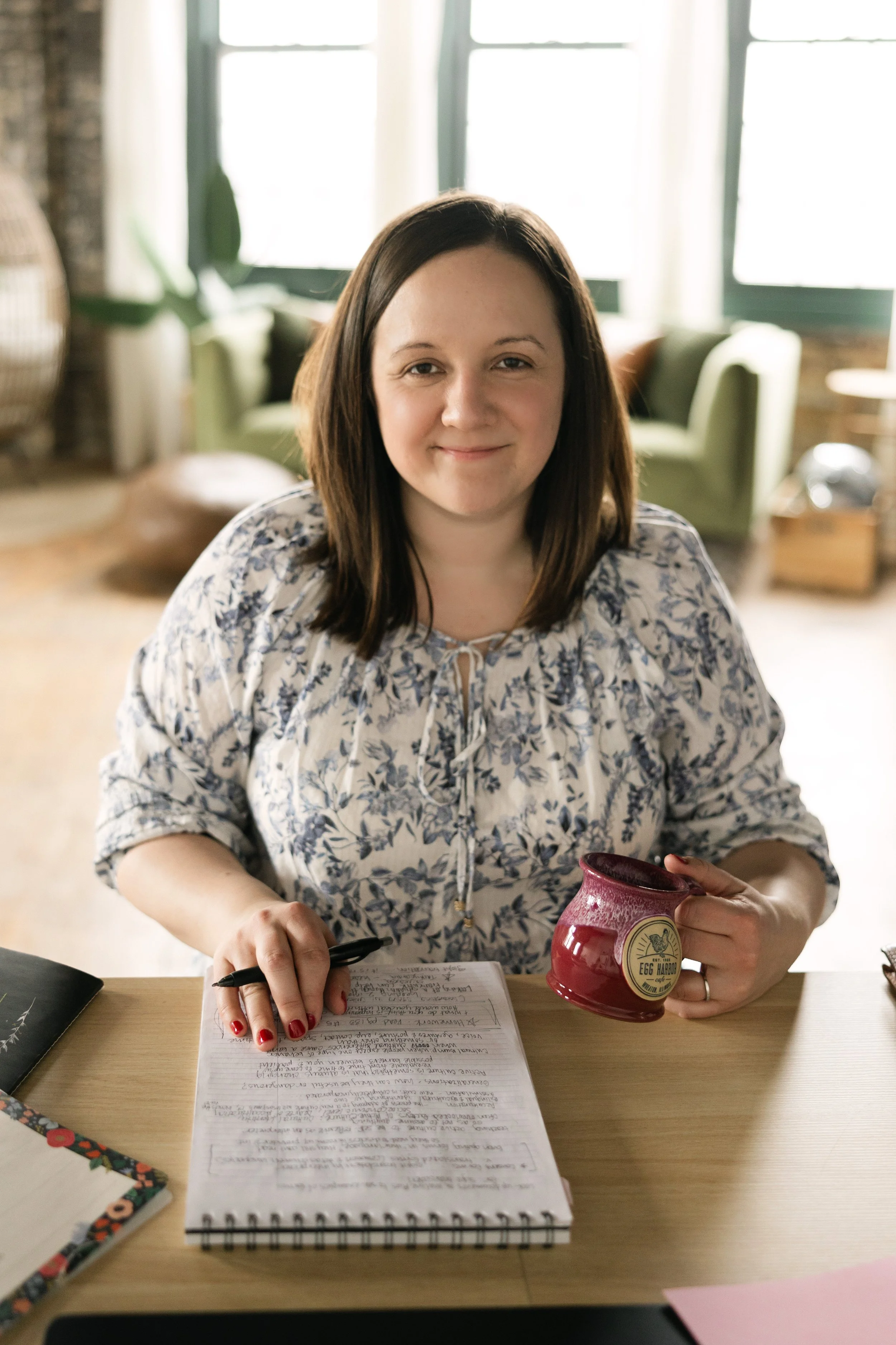 Rachel smiles at the camera holding a coffee mug and a pen with a notebook in front of her