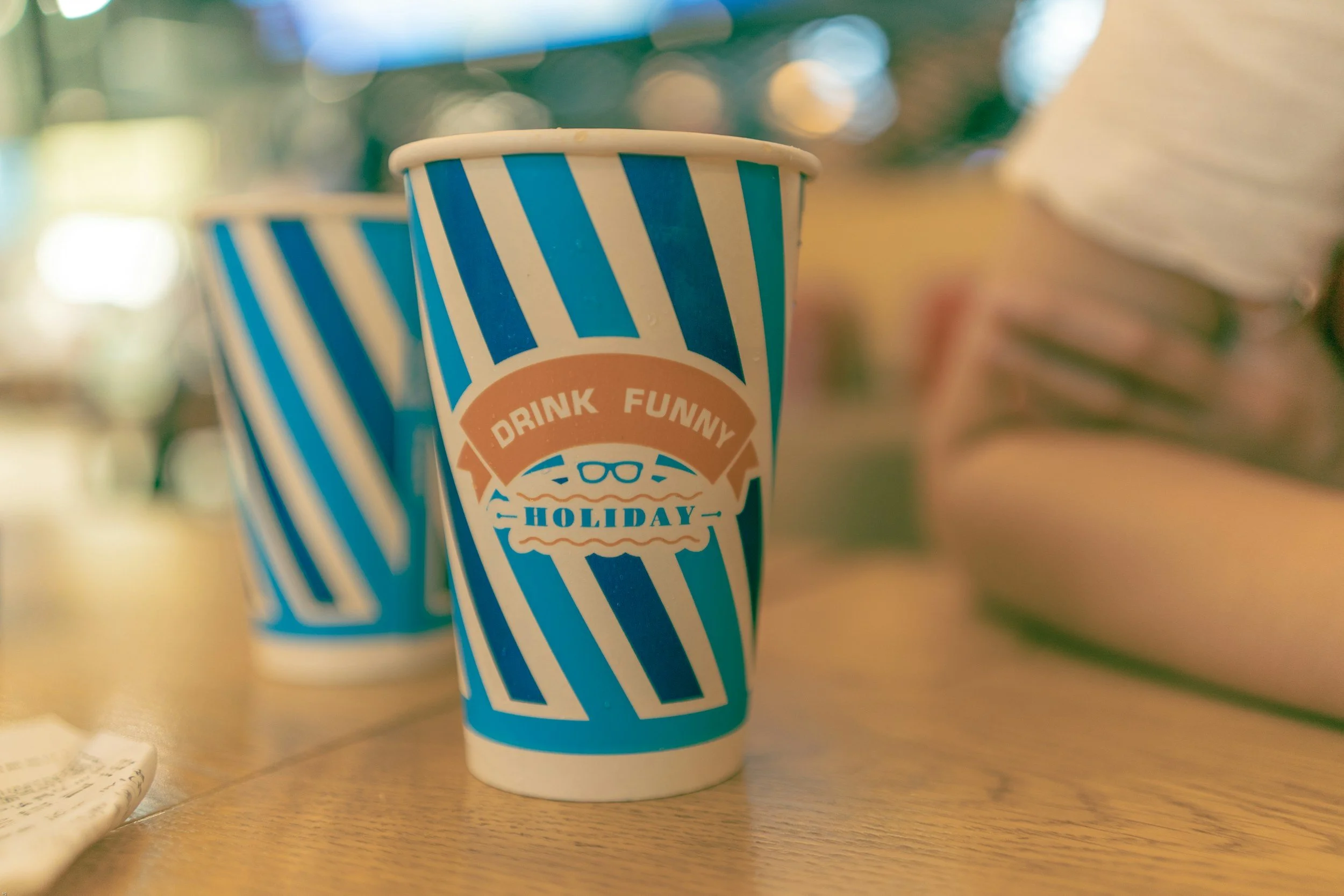 Two blue and white striped paper soda cups on a wooden table, one in the foreground with a label that says "Drink Funny Holiday" with glasses and a burger illustration.
