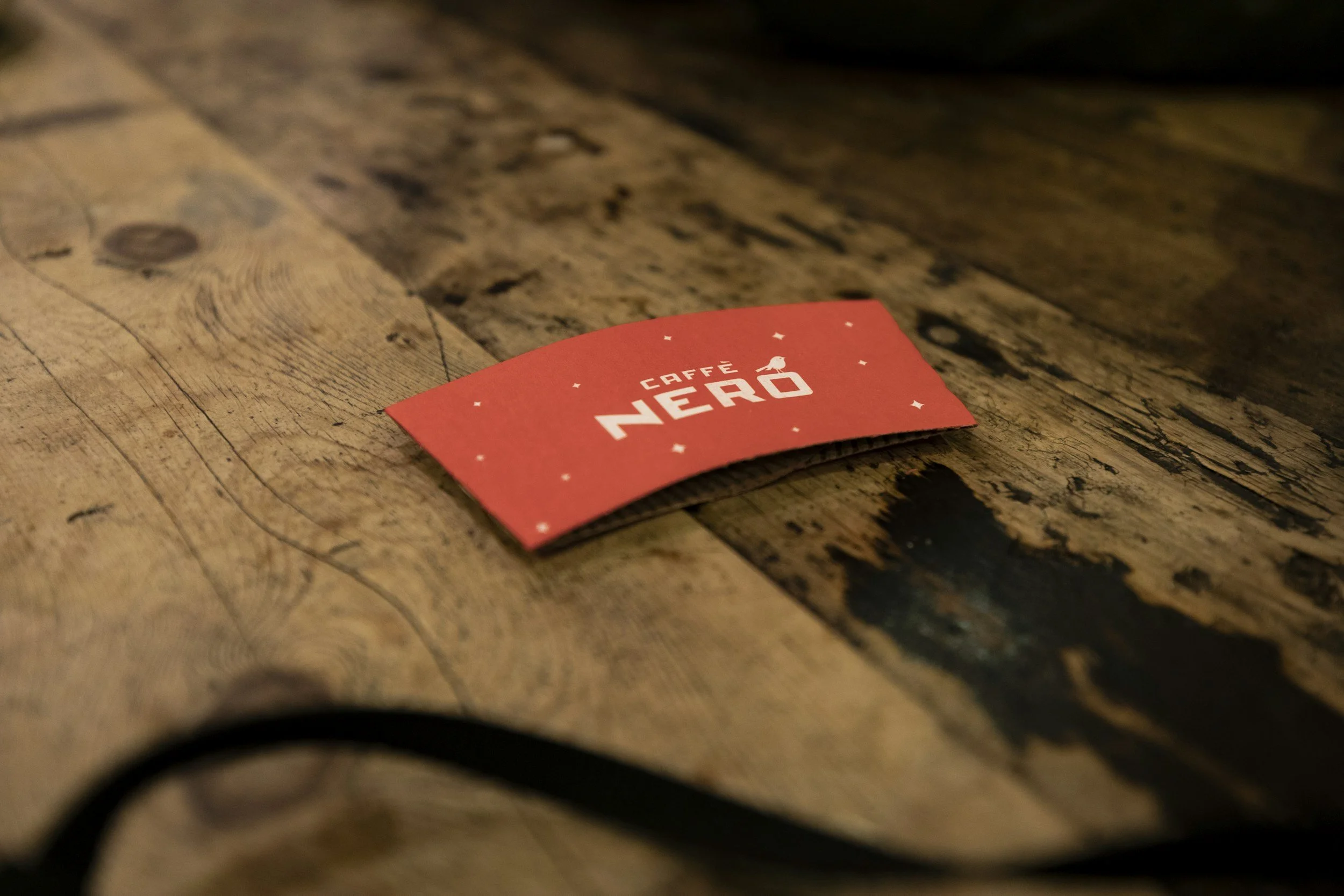 A red coffee cup sleeve with the words 'Caffé Nero' and a small bird icon, lying on a rustic wooden table.
