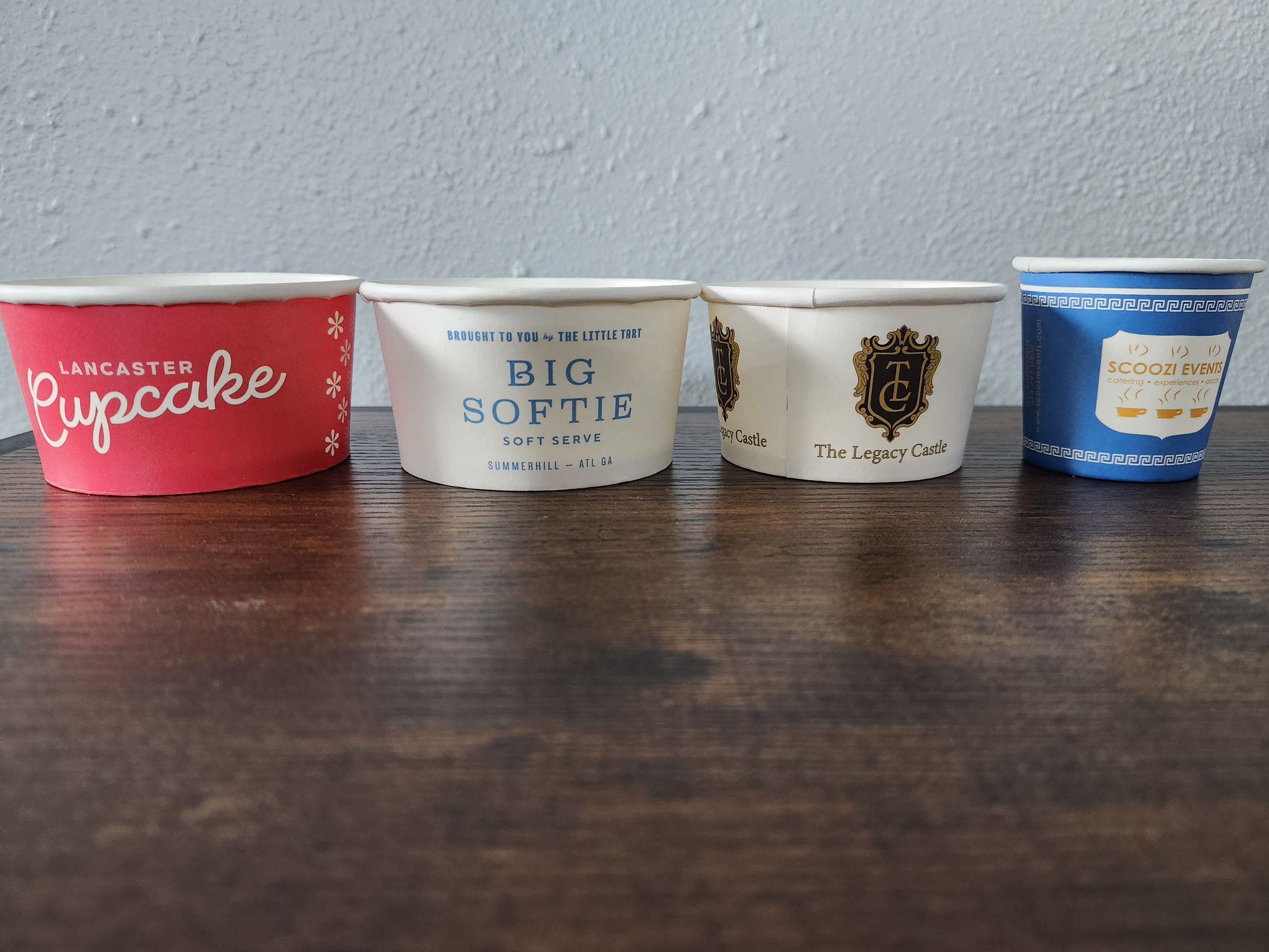 Four small paper food and ice cream bowls with different branding, placed on a wooden surface against a plain wall background.