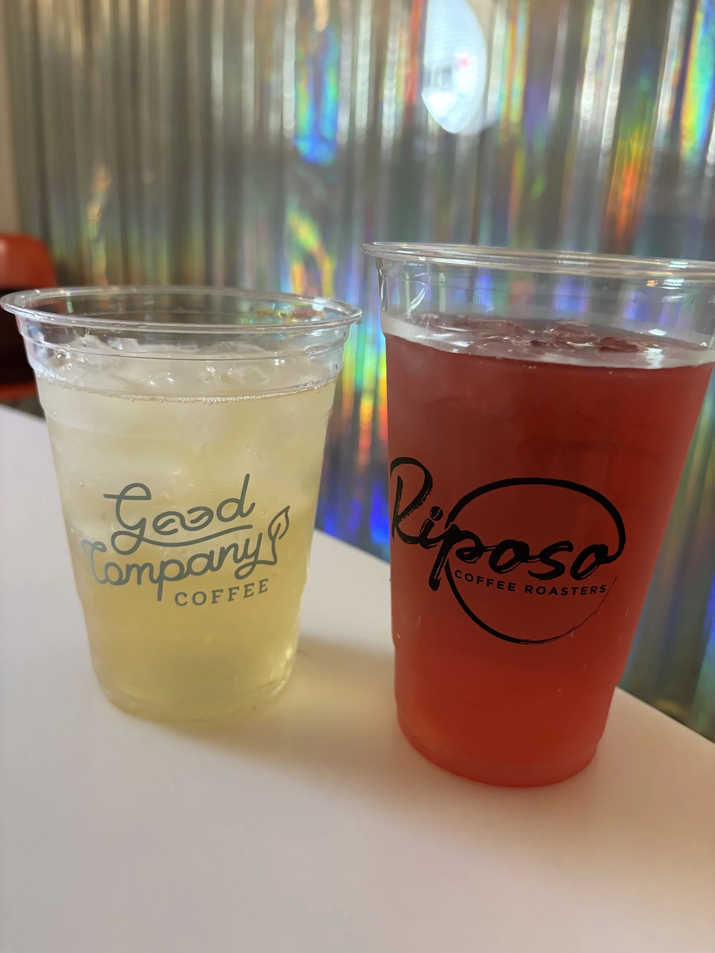 Two cups of beverages on a white surface, one with a light yellow drink labeled 'Good Company Coffee' and the other with a red-colored drink labeled 'Riposo Coffee Roasters', against a multicolored, holographic background.