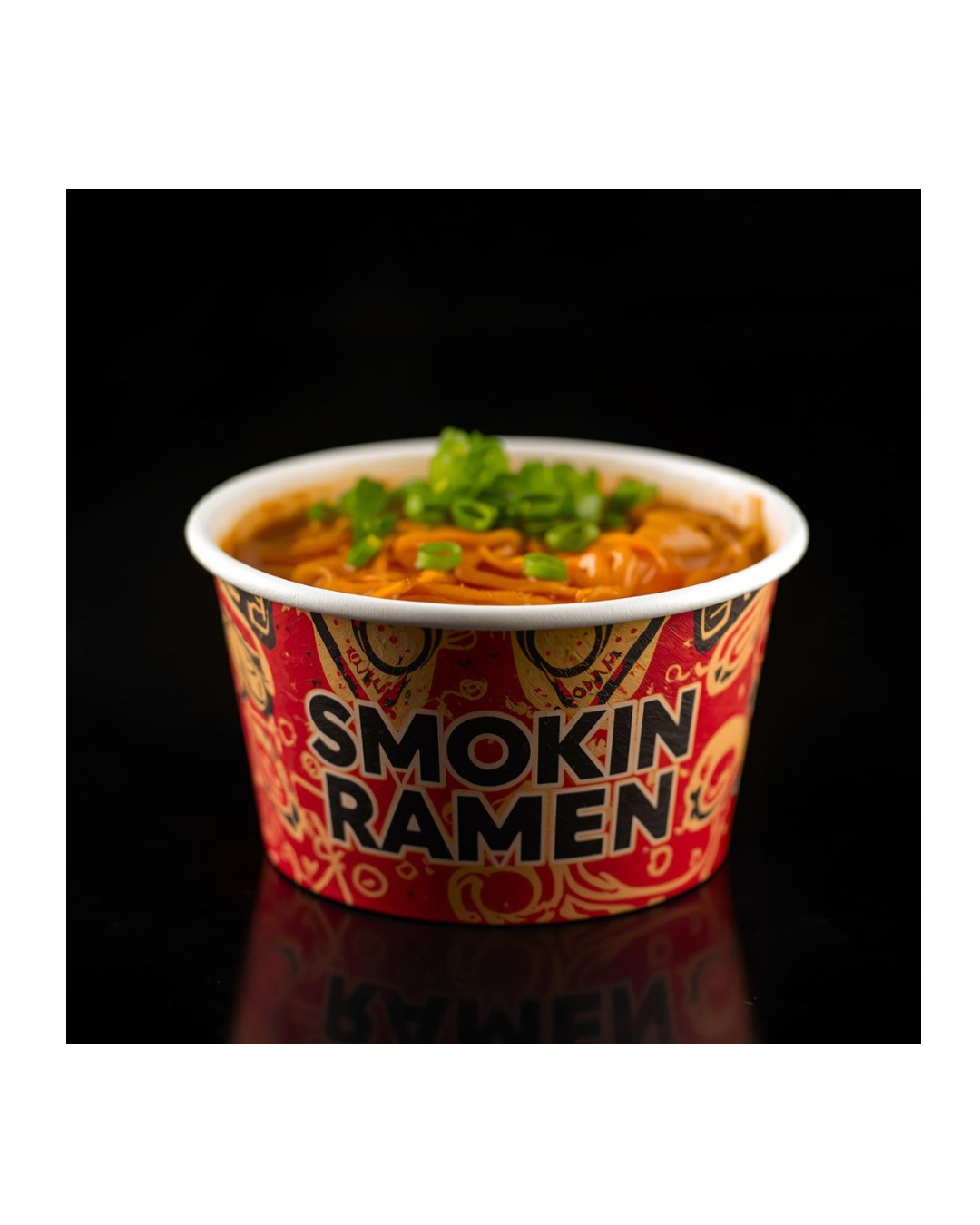 A red and black paper food container labeled "Smoking Ramen" filled with ramen noodles topped with chopped green onions, against a black background.