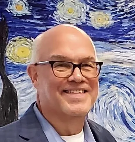a man wearing glasses in a suit smiles in front of artwork.