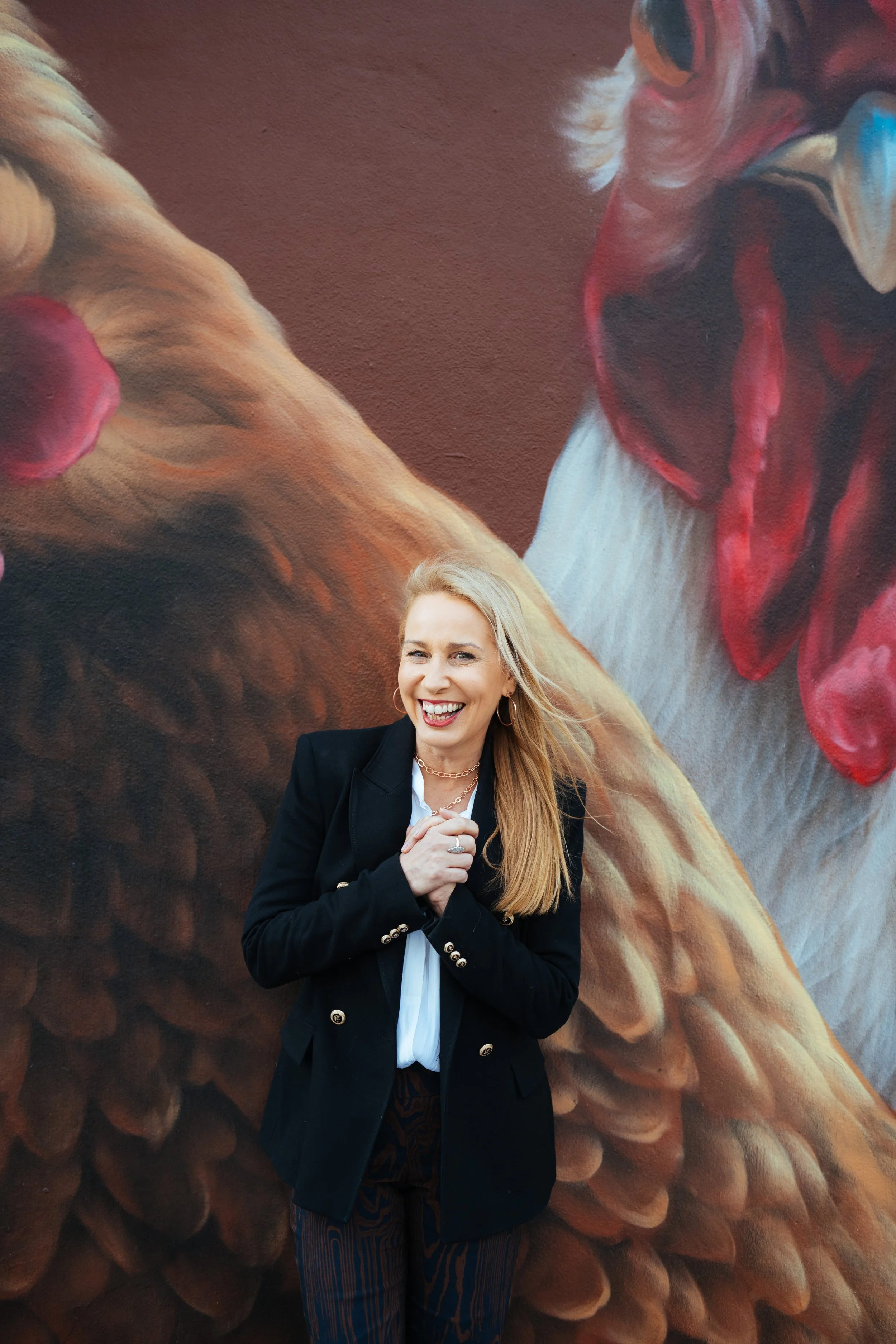 A woman with long blonde hair smiling and wearing a black blazer with gold buttons, a white shirt, and striped pants, standing in front of a detailed mural of a chicken.